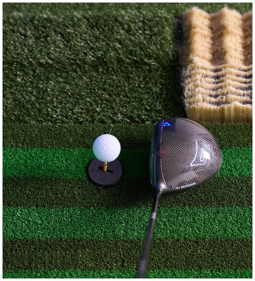 A wood golf club laying behind a ball teed up on fake grass. Beside is another piece of fake grass with brush-like brown bristles to simulate a sand trap