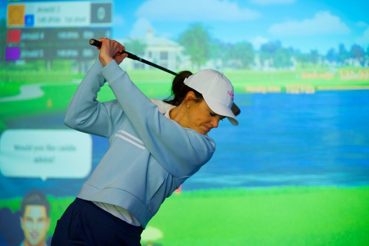A woman in a white golf hat and blue shirt takes a swing in front of a golf simulator