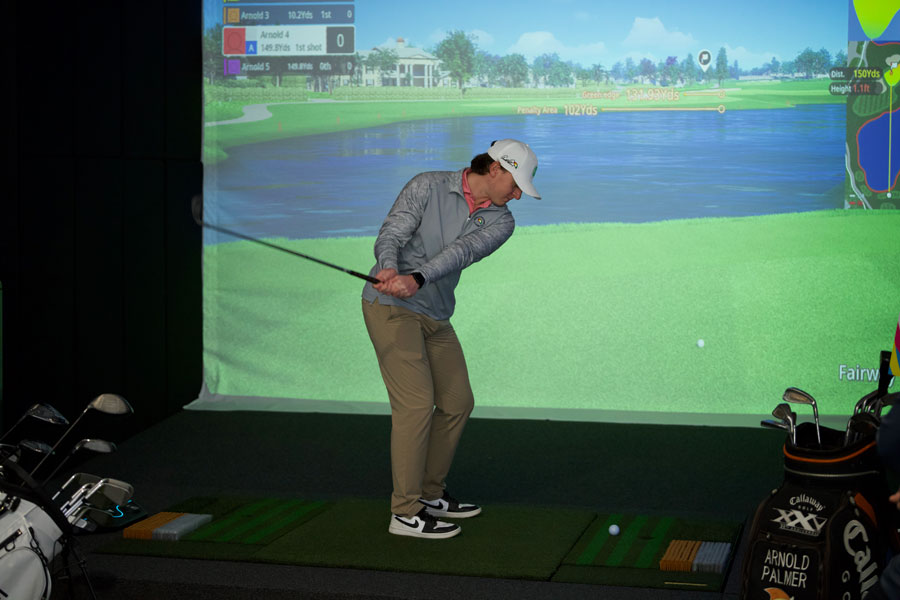 A man in golf attire in the middle of his backswing. The golf simulator screen is showing a large pond in the middle of the fairway.