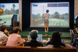 Four young men are seated on a wooden bench watching the four finish taking a shot on the golf simulator.