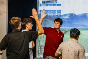Four young men gathered around the golf simulator wearing golf shirts. Two of them are high-fiving.