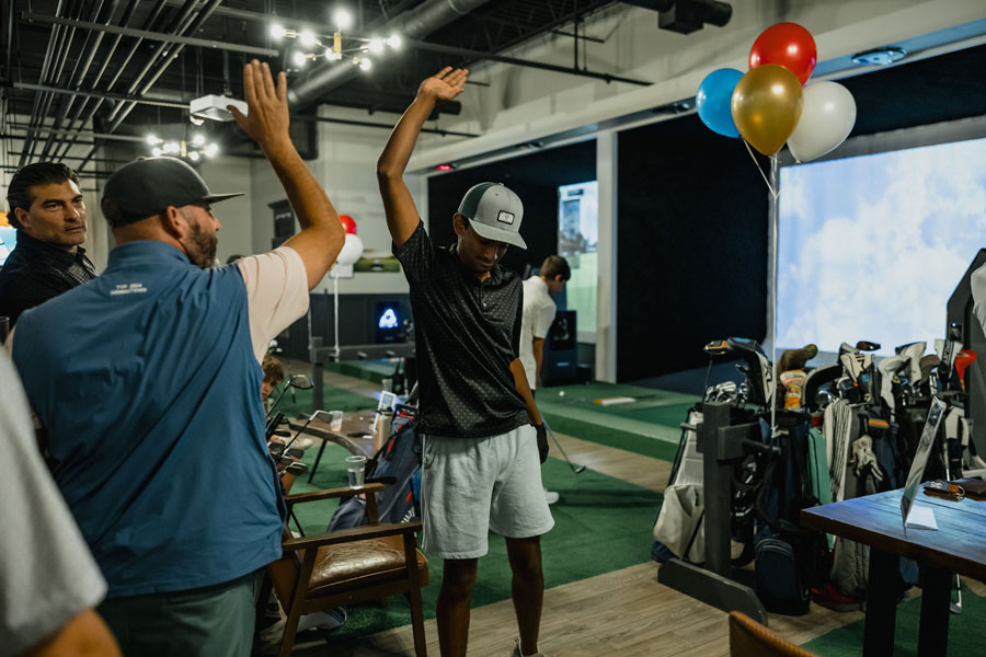 Two men and a teen boy are gathered at a golf simulator. One of the men and the boy have one arm raised in celebration.