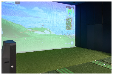 Fake green grass on the floor, one with a golf ball on it. The wall in front has an image showing the tee off spot and with green fairway and sand bunkers in the distance